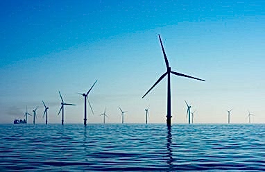 A wind farm out at sea shows around 6 turbines in the foreground with more in the far distance under a bright blue sky.