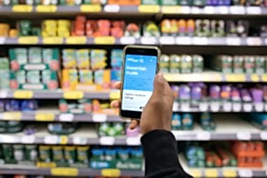 A hand holding up a phone with Marine Conservation Society's Good Fish Guide app on the screen. In the background is a blurred supermarket shelf filled with food products.
