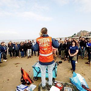A woman wearing a hi-vis orange vest has her back to the camera. She is in front of a group of volunteers. Her vest reads Marine Conservation Society.