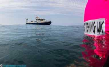 A fishing boat in shown in the distance with a bright pink buoy-type device reading 'ghost fishing UK' in the foreground.