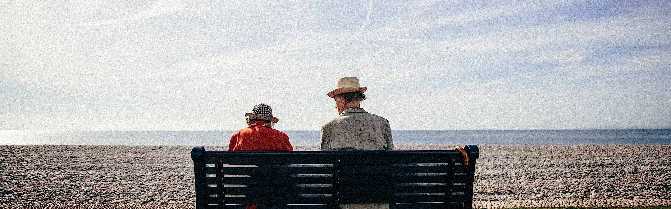 An elderly couple sitting on a dark green bench wth their backs to the camera. They are looking over a shingle beach toward a calm, blue sea. The man has a light coloured blazer and straw hat and the woman wears a red jacket and patterned bucket hat.