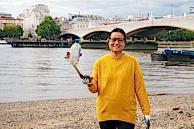 A woman in a yellow jumper holding up a plastic bottle to the camera using a litter picker. She is in front of the river at Festival Pier, London