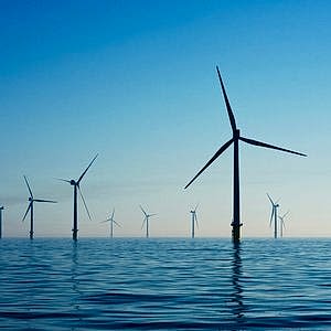 A wind farm out at sea shows around 6 turbines in the foreground with more in the far distance under a bright blue sky.