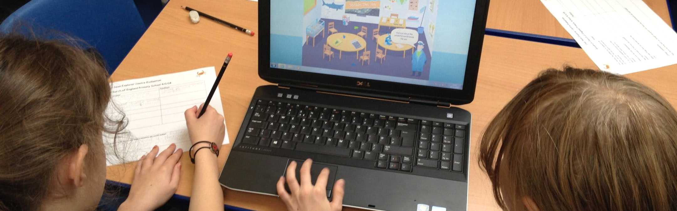The back of 2 primary school girls' heads looking at a laptop screen on a table. The laptop screen shows a cartoon sea captain standing iinside a classroom.