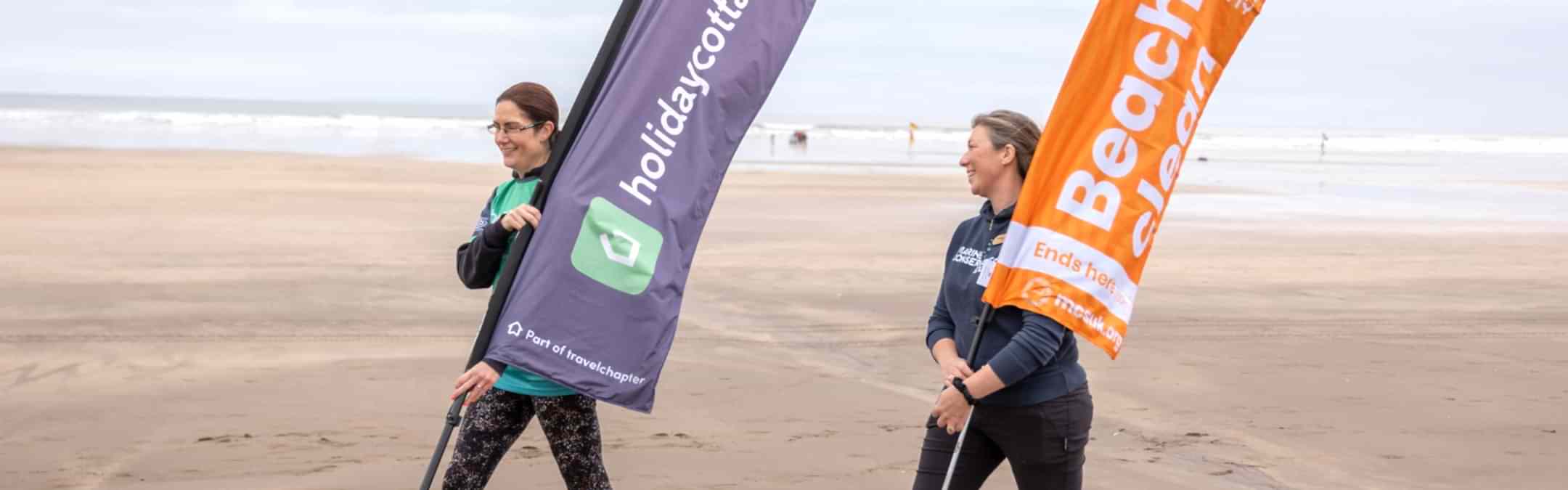 2 women walking along the beach carrying large marker flags. The flags read "Holidaycottages.co.uk" and "Marine Conservation Society Beach Clean" respectively