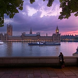 Westminster at sunset taken from the bank across the Thames. The sky is a stunning purple pink and an ornate lamp is lit in the foreground.