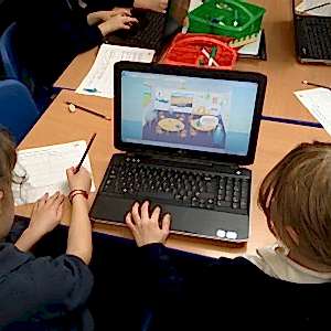 The back of 2 primary school girls' heads looking at a laptop screen on a table. The laptop screen shows a cartoon sea captain standing iinside a classroom.