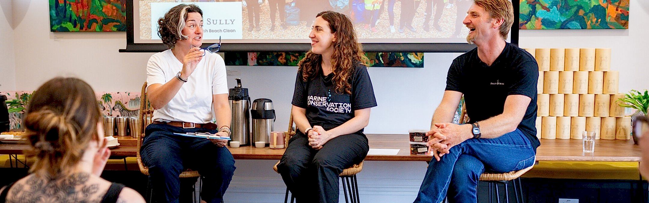 Zoe Lyons, a Marine Conservation Society staff member, and Cully from Cully and Sully, sit on stools in front of a large screen, giving a talk to an audience. All three people are smiling broadly.