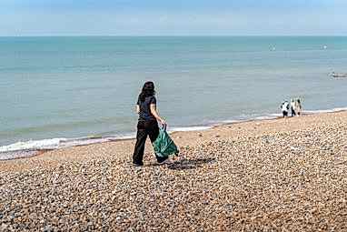 A beach cleaner walks with her back to the camera on a rocky UK beach with the sea to her left.. From the clear blue sky and her sleeveless clothing, it looks like a beautiful UK summer day.