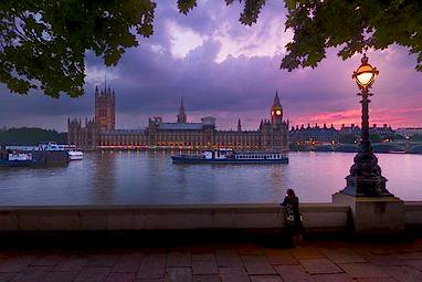 Westminster at sunset taken from the bank across the Thames. The sky is a stunning purple pink and an ornate lamp is lit in the foreground.