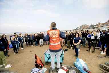 A woman wearing a hi-vis orange vest has her back to the camera. She is in front of a group of volunteers. Her vest reads Marine Conservation Society.