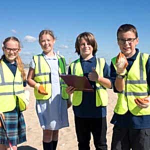 4 school children looking at the camera wearing hi-vis jackets and gloves on. The girl on the far left is holding a litter picker and the boy that is centre right is holding a clipboard. They are on a beach