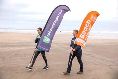 2 women walking along the beach carrying large marker flags. The flags read "Holidaycottages.co.uk" and "Marine Conservation Society Beach Clean" respectively