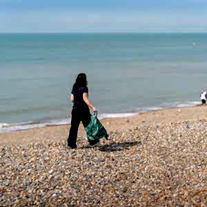 A beach cleaner walks with her back to the camera on a rocky UK beach with the sea to her left.. From the clear blue sky and her sleeveless clothing, it looks like a beautiful UK summer day.