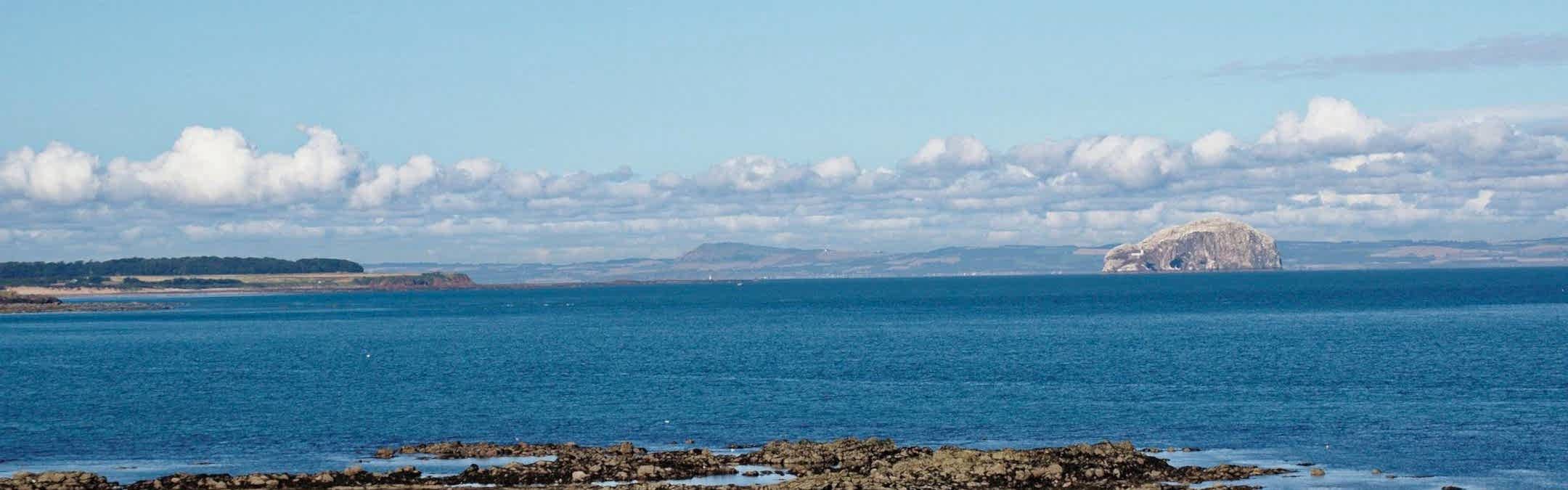 A view out over the Firth of Forth with a rocky shore in the foreground and a beautiful view across the water. The sky is blue and the clouds sit low in the sky.